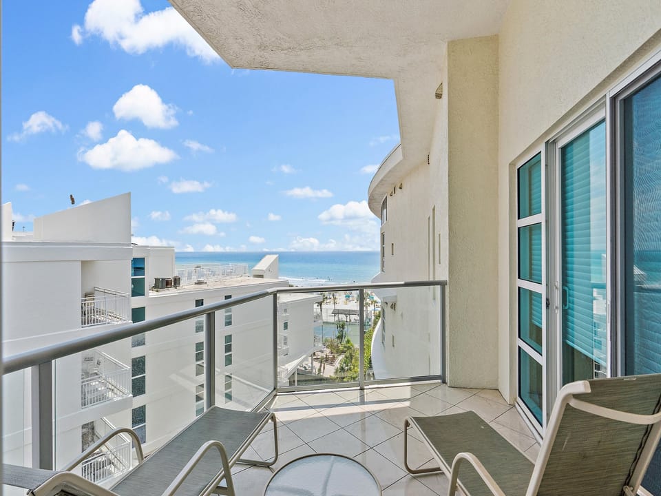 A balcony with a glass railing features several lounge chairs and a small table. The view overlooks modern white buildings and extends to the ocean under a blue sky with scattered clouds.