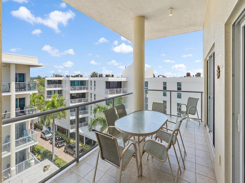 A spacious balcony with a glass-top table and six chairs overlooks modern apartment buildings and trees under a blue sky with scattered clouds.