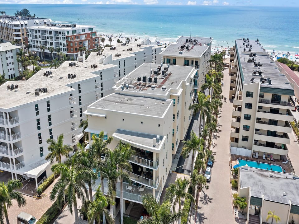 Aerial view of beachfront condos with palm trees, a swimming pool, and the ocean in the background. People are on the beach and in the water. The image shows a sunny day with some clouds in the sky.