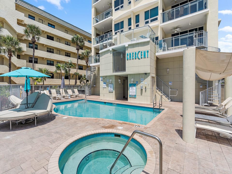 Outdoor pool area beside a multi-story building with lounge chairs, umbrellas, and a hot tub. The building has balconies with glass railings and a sign that reads "Crescent.