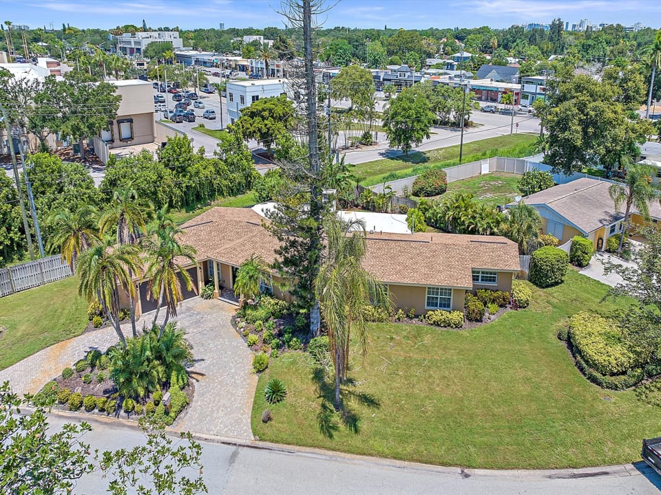 An aerial view of a single-story home with a brown roof, surrounded by a fenced yard and greenery, located at a street intersection within a suburban area with both residential and industrial buildings.