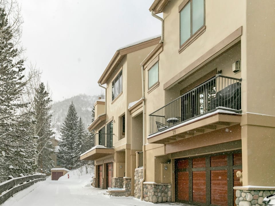 A row of beige townhouses with garages and balconies, set alongside a snow-covered pathway bordered by a wooden fence, surrounded by trees and hills.
