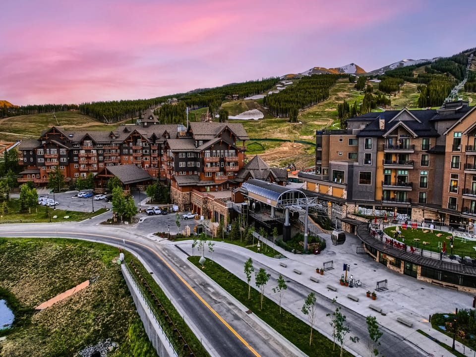 Aerial view of a mountain resort with large buildings, a winding road, and ski lifts under a pink and purple sunset sky, with green slopes in the background.