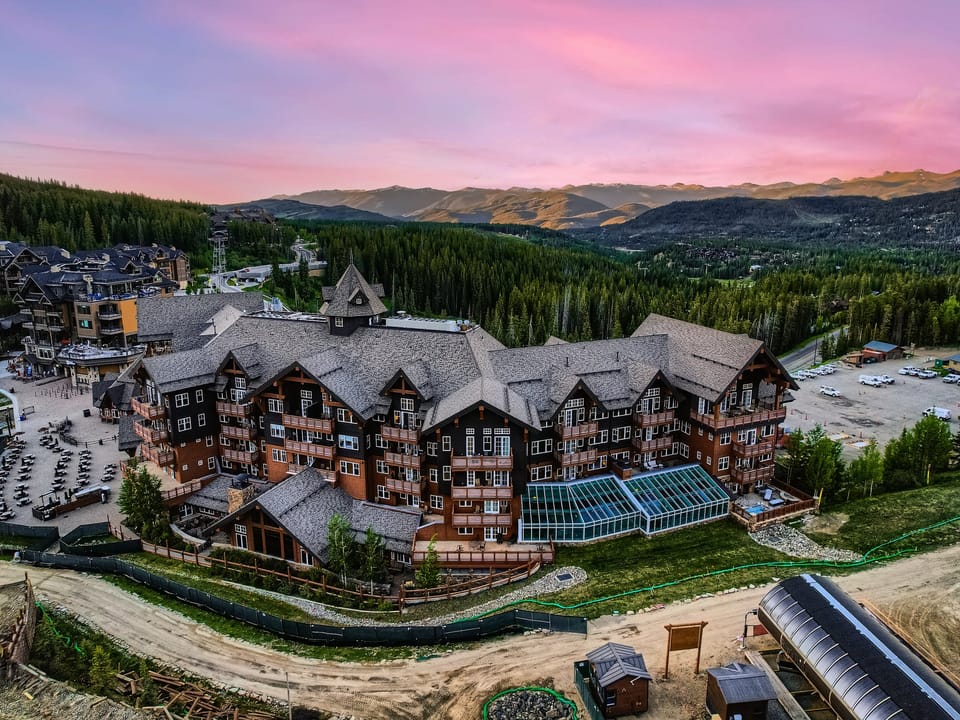 A large lodge-style resort with multiple buildings surrounded by forested hills, against a backdrop of mountains under a pink and purple sunset sky.