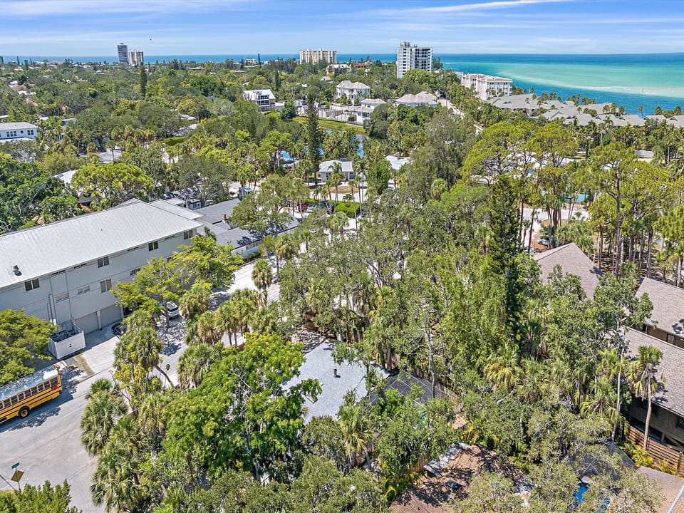 Aerial view of a coastal suburban neighborhood with numerous trees, white buildings, a yellow school bus, and the ocean visible in the background.