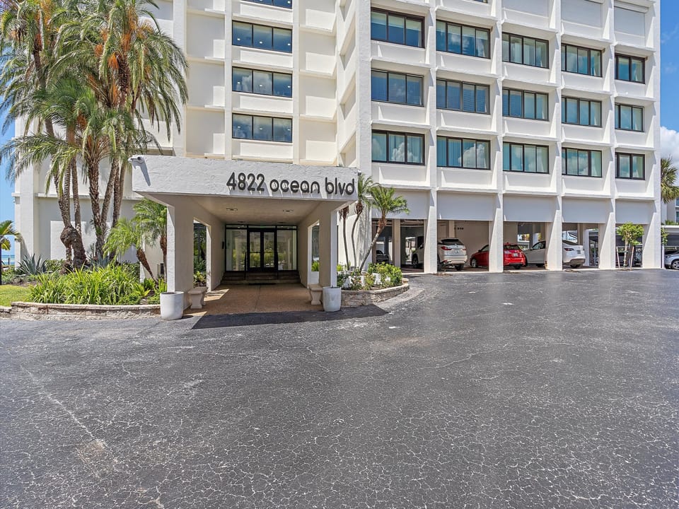 A white building entrance with the address "4822 Ocean Blvd" displayed above. Several floors with windows are visible, flanked by palm trees. Cars are partially visible in a parking area to the right.
