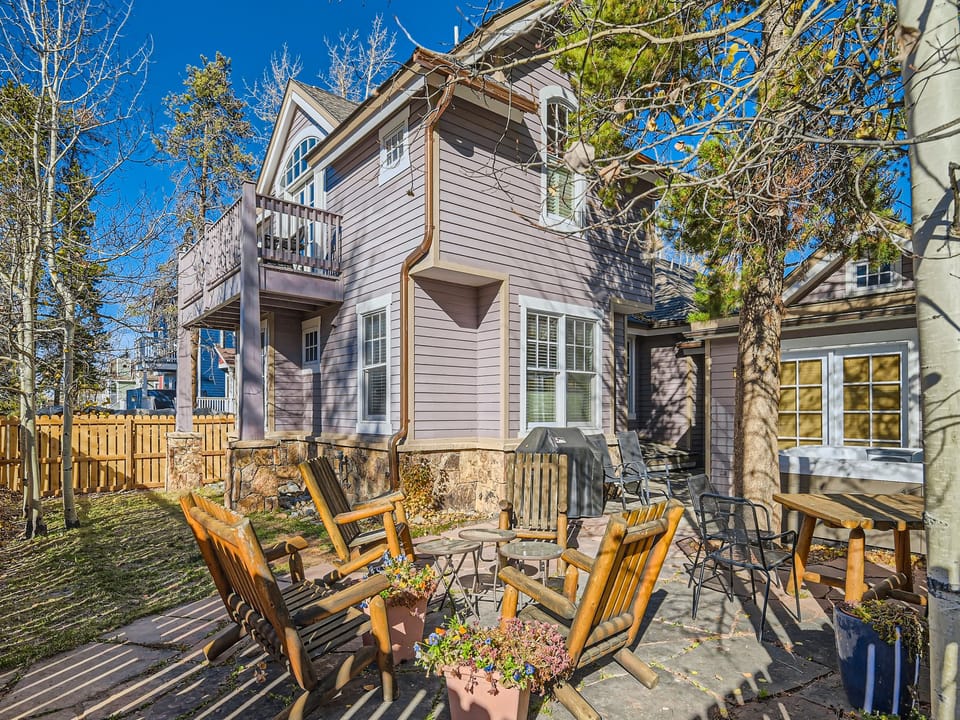 A two-story light purple house with outdoor seating and a small garden area. The backyard features wooden chairs surrounding a fire pit.