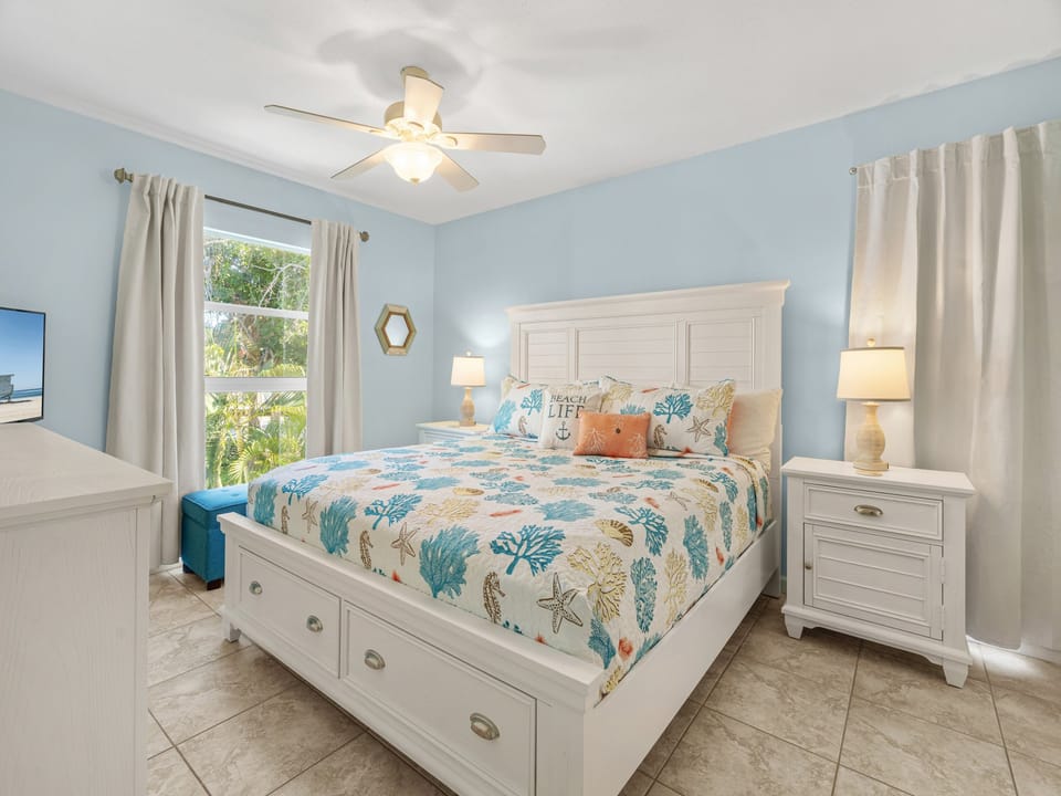 Your coastal bedroom features serene blue walls, ocean-themed bedding, and garden views through bright windows—perfect for peaceful mornings.