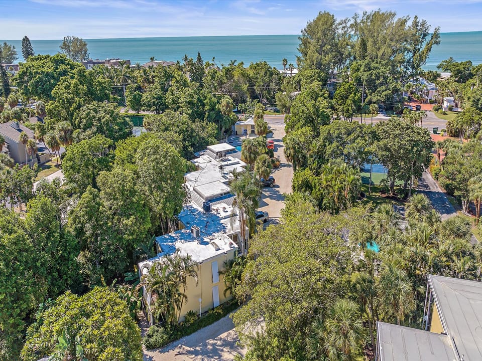 Aerial view of a coastal residential area with multiple buildings surrounded by trees and vegetation, with the ocean visible in the background on a sunny day.