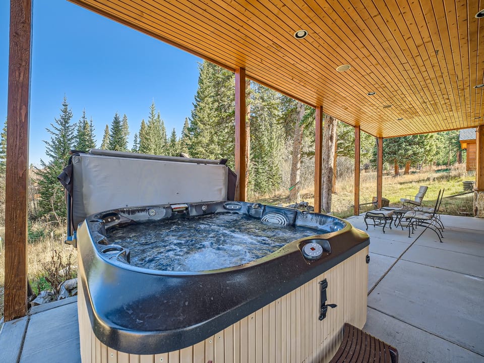 A hot tub under a wooden patio cover with trees and outdoor furniture in the background.
