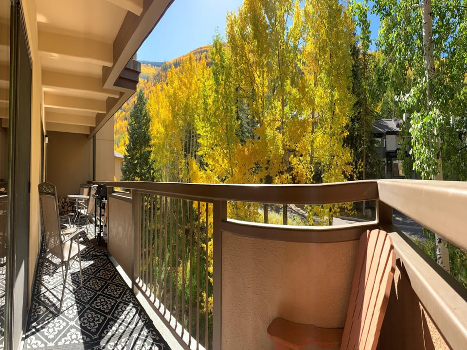 A balcony with chairs offers a view of trees with autumn foliage. The floor has a decorative black-and-white pattern. Nearby houses are partially visible through the trees.