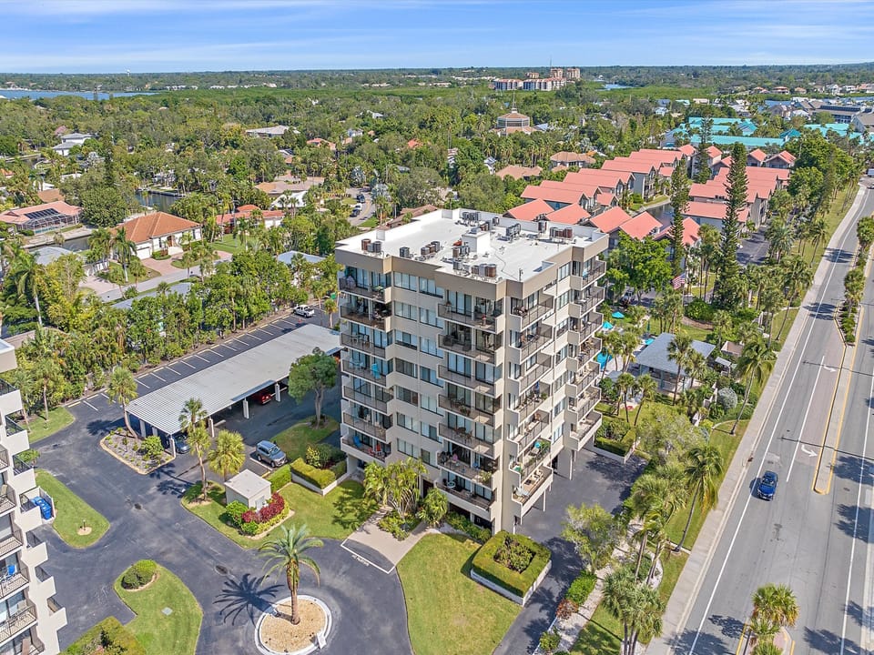 Aerial view of a mid-rise residential building surrounded by lush greenery, with a parking area and a main road adjacent to the property.