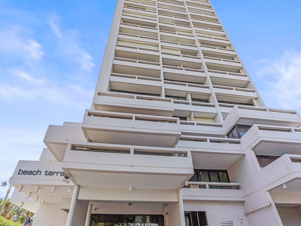 A tall, modern apartment building named "Beach Terrace" with multiple balconies, viewed from a low angle against a blue sky.