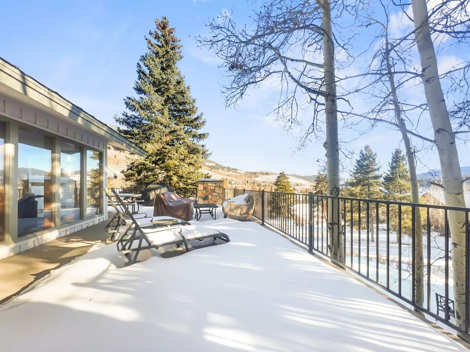 Outdoor deck covered in snow, furnished with lounge chairs and patio furniture, overlooks a scenic winter landscape with trees and distant hills under a clear blue sky.