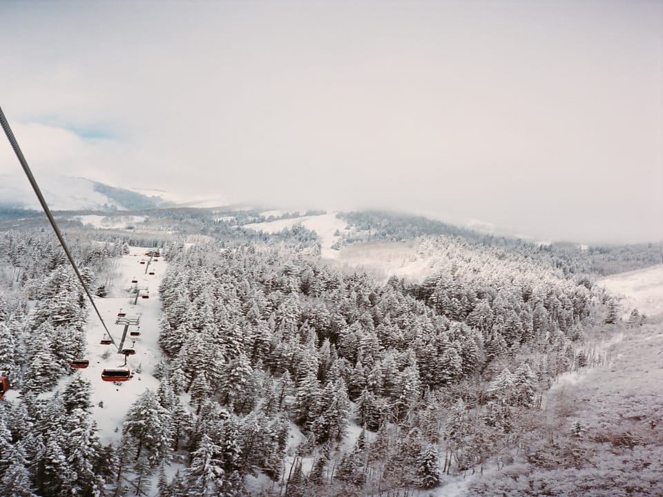 An aerial view of a snow-covered forest with a ski lift ascending into the foggy distance, surrounded by mountainous terrain.