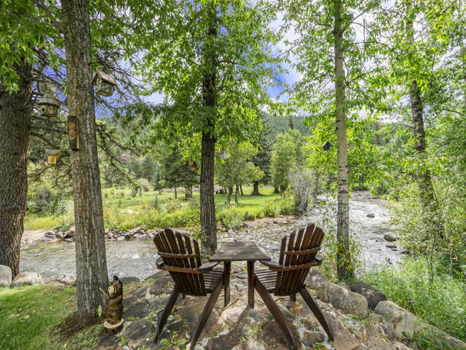 Elk River Cabin -- Cabin Condo in Estes Park -- 23-ZONE3554 - a SkyRun RMNP Property - Elk River Cabin - Elk River Cabin Adirondack chairs on riverbank in backyard.