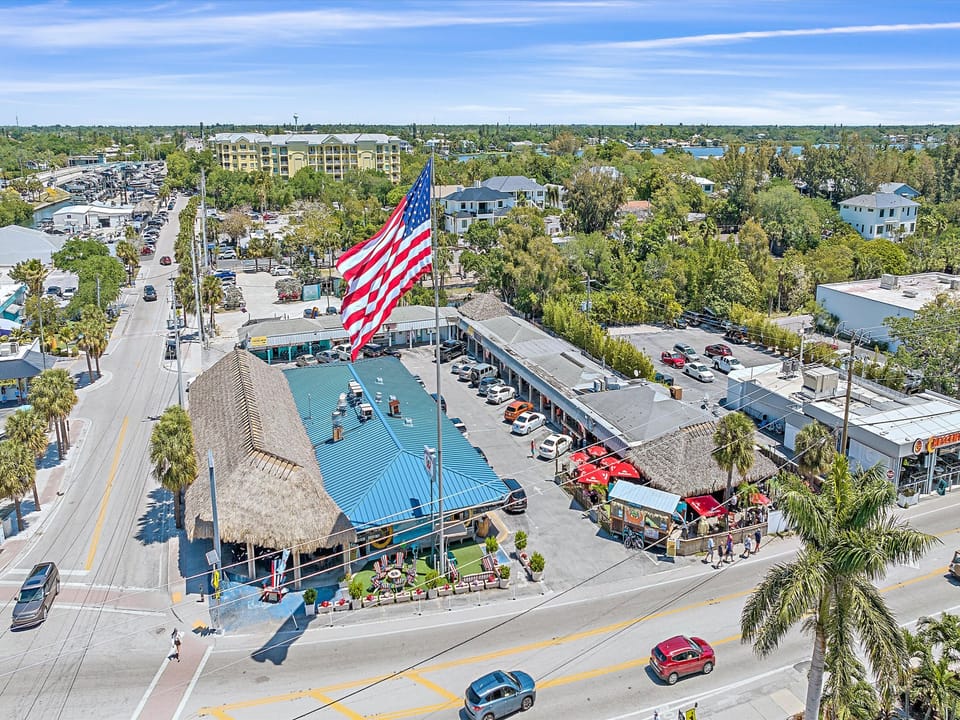 Aerial view of a commercial area with buildings, parking lot full of cars, and an American flag prominently displayed. Roads, trees, and other buildings are visible in the background.