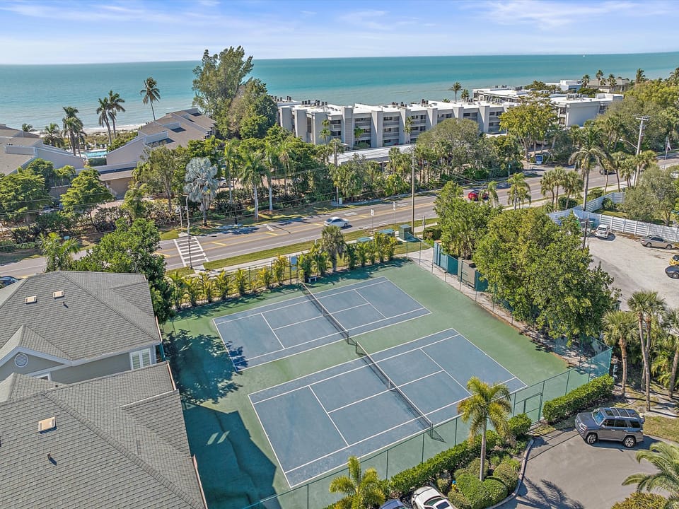 Aerial view of a tennis court in a coastal area, surrounded by buildings, palm trees, and a road with ocean view in the background.