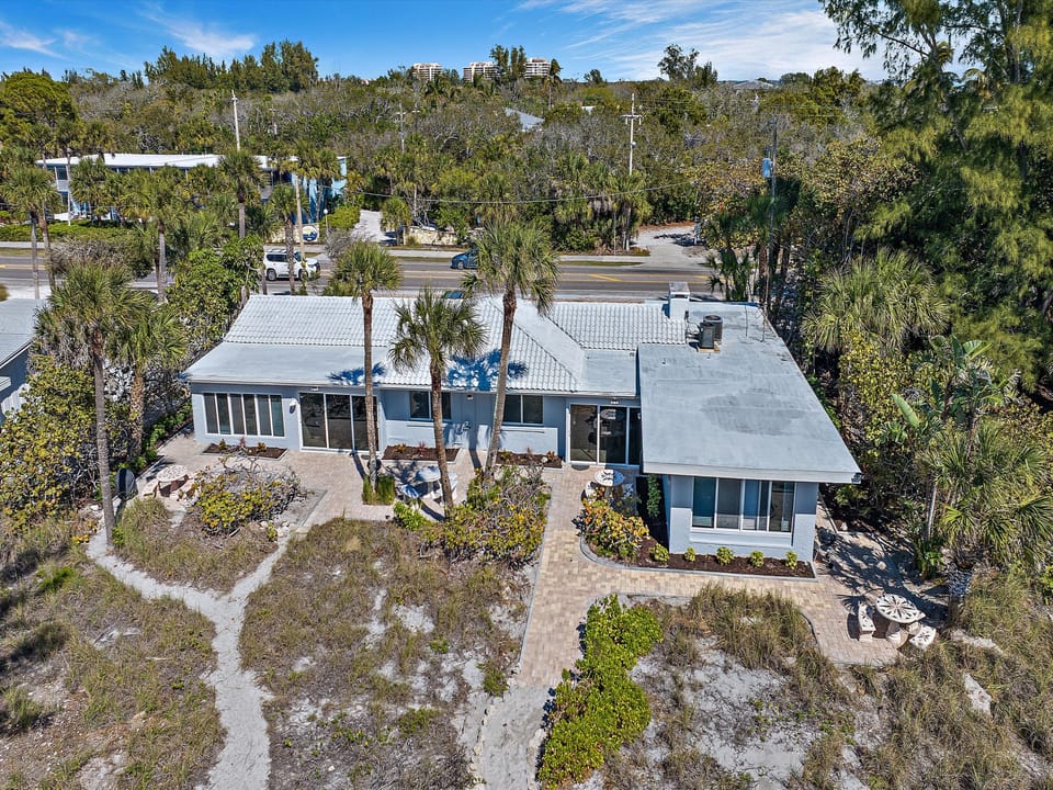 Aerial view of a single-story house with blue trim, surrounded by vegetation and featuring a stone pathway, located near a road with additional buildings in the background.
