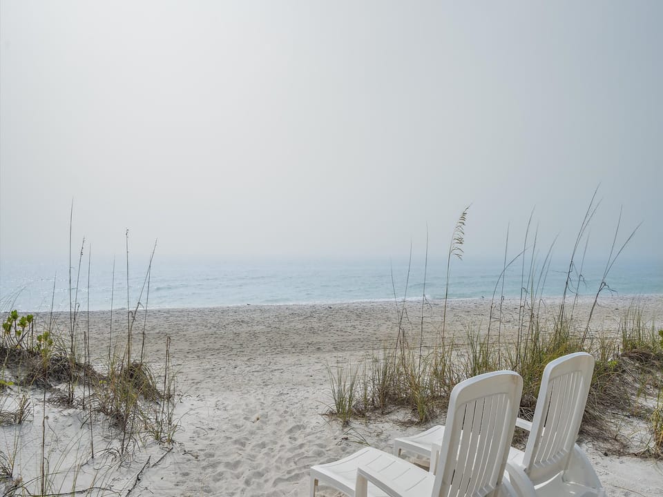 Two white lounge chairs facing a misty, empty beach with calm ocean waves and sparse vegetation in the foreground.