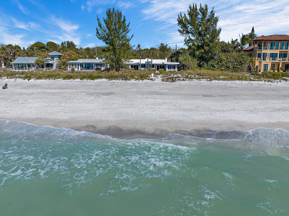 A beachfront view shows gentle waves reaching a sandy shore lined with trees and buildings under a partly cloudy sky.