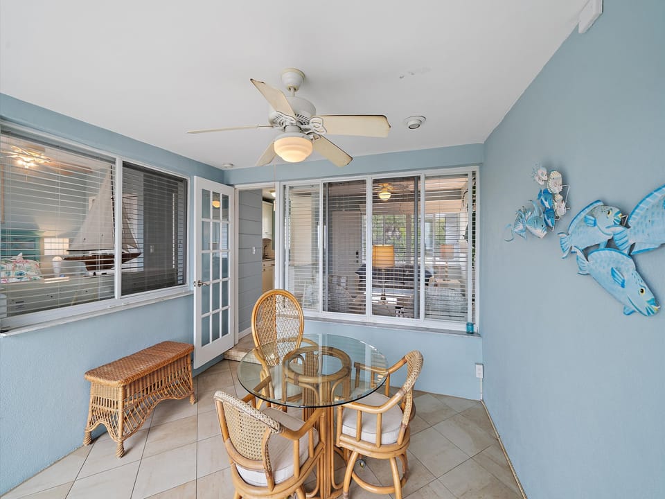 A sunroom with light blue walls featuring a glass-top table with four wicker chairs, a wicker bench, a ceiling fan, and decorative fish on the wall. Windows and a door provide a view of another room.
