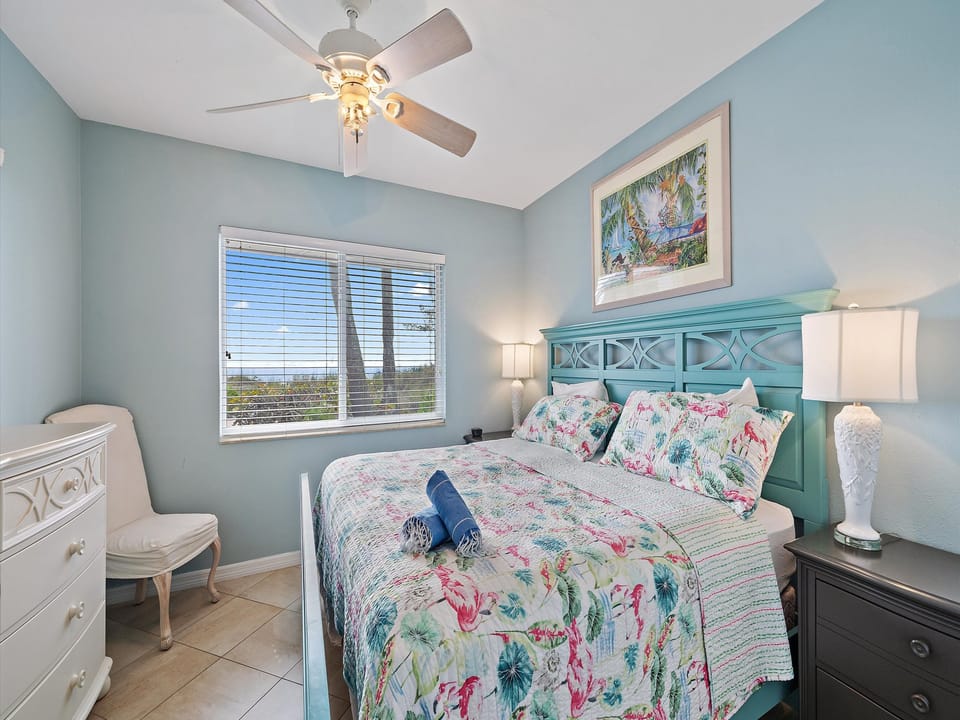 A bedroom with a king-size bed featuring floral bedding, and flanked by two bedside tables with lamps. There is a ceiling fan, a chest of drawers on the left, and a window with outdoor views.