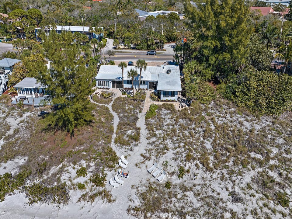 Aerial view of a beachfront property with two white-roofed buildings, pathways leading to beach chairs, and a road with cars in the background.