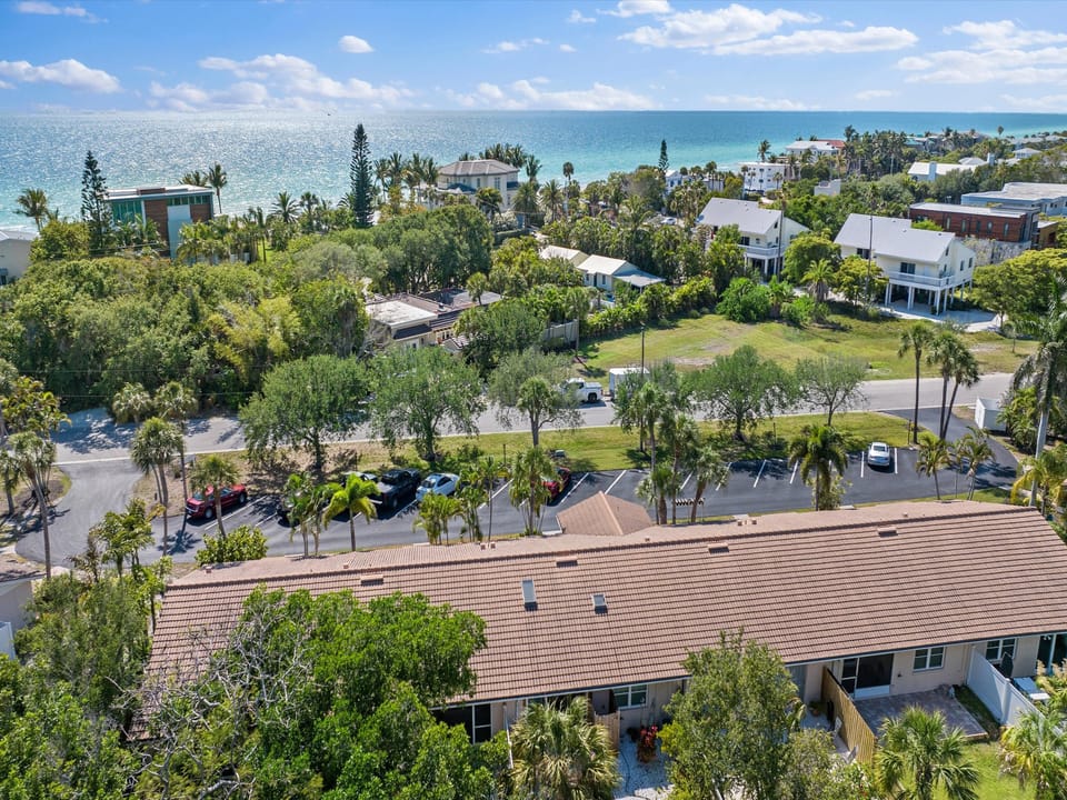 Aerial view of a coastal neighborhood with houses, trees, parked cars, and a nearby ocean on a clear day.