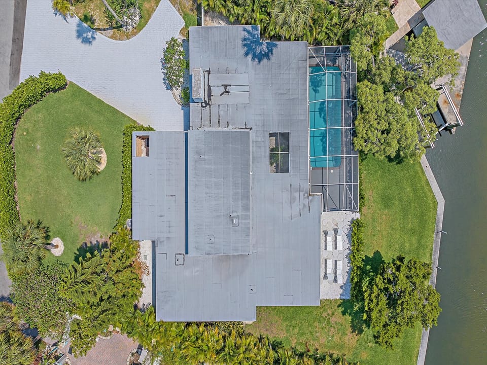 Aerial view of a modern house with a grey roof, landscaped garden, and a screened-in swimming pool adjacent to a body of water.