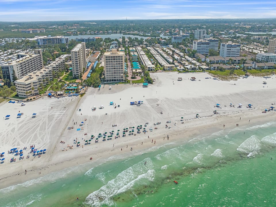 Aerial view of a sandy beach with people and umbrellas near the shore, adjacent to a row of tall buildings and residential areas. Waves crash gently against the shoreline.