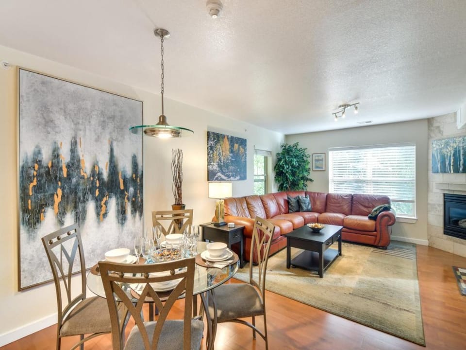 A cozy living room with a brown leather sectional sofa, a dining table set for four, and contemporary art on the walls. The room has wooden flooring and large windows allowing natural light.