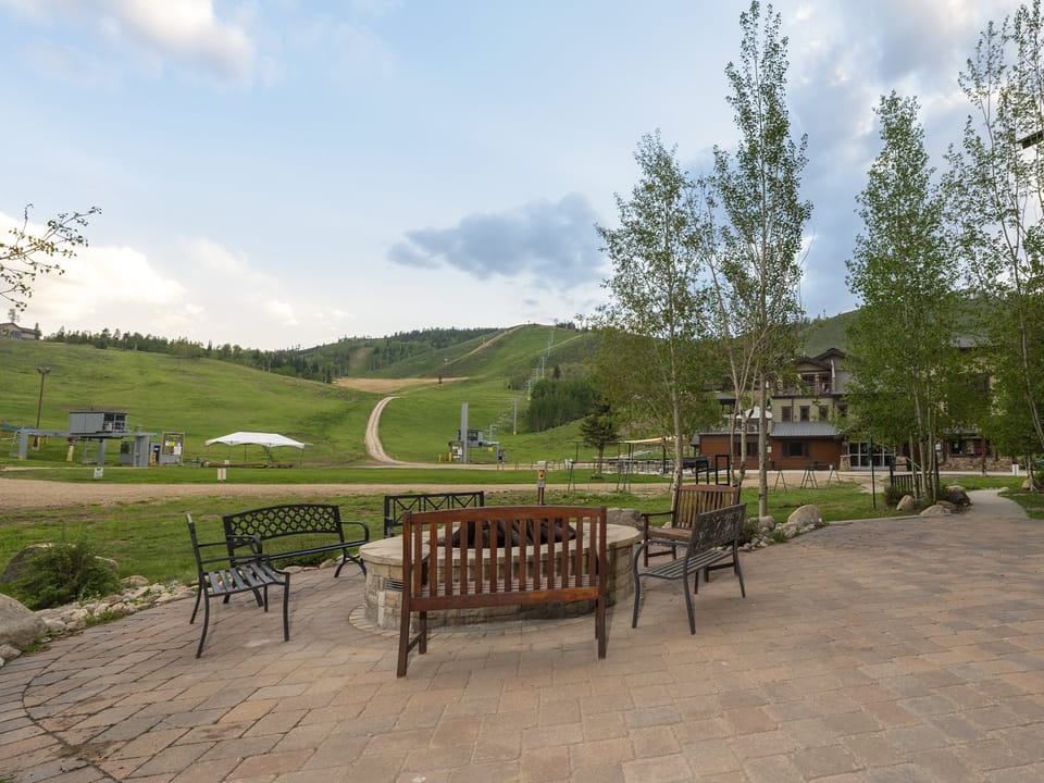 Shared outdoor patio with a fire pit, benches, and chairs on a paved patio, surrounded by trees