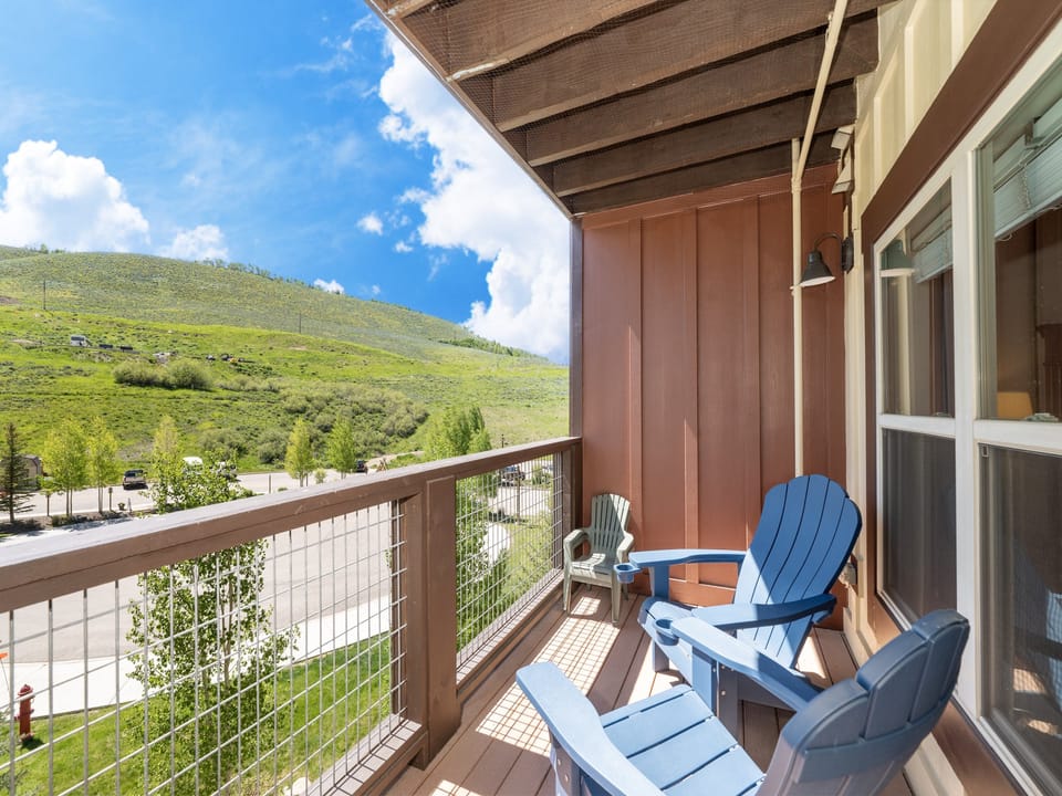 A balcony with blue Adirondack chairs overlooks a green hillside on a sunny day.