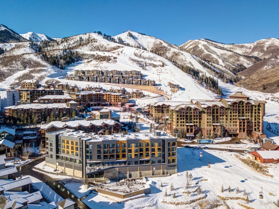 A snow-covered resort area with multiple modern buildings and residences nestled in a mountainous region under a clear blue sky.