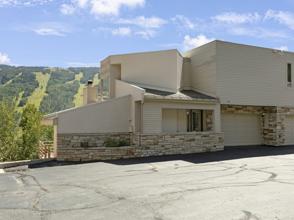Modern house with a flat roof and stone accents, set against a backdrop of green hills and blue sky.