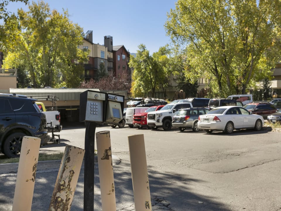 Parking lot with various vehicles under clear blue sky; foreground features parking meter and yellow barriers. Trees and buildings in the background.