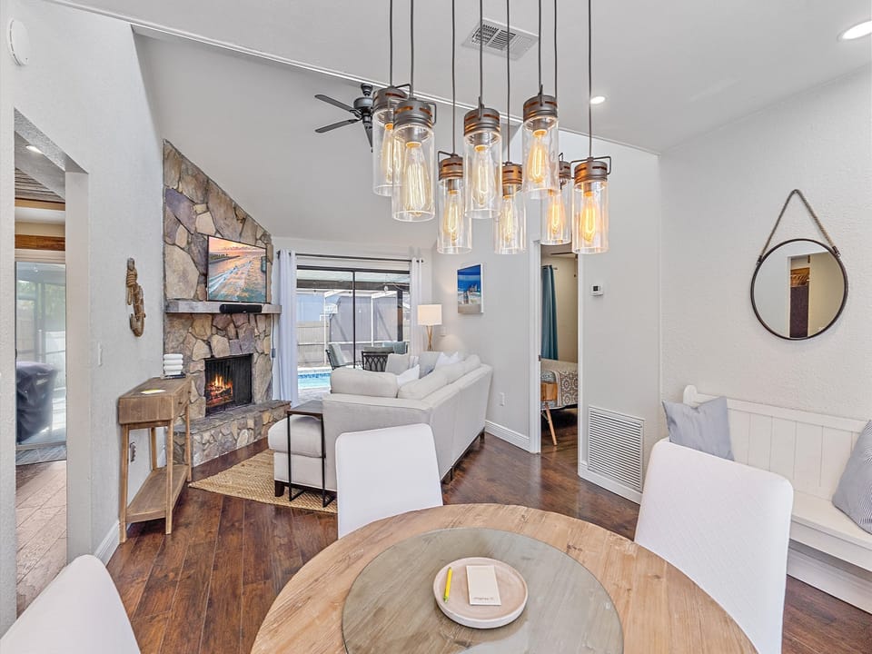 A modern living room with a stone fireplace, flat-screen TV, sectional sofa, and glass doors opening to a pool. A round dining table with a book and a chandelier hanging overhead is in the foreground.