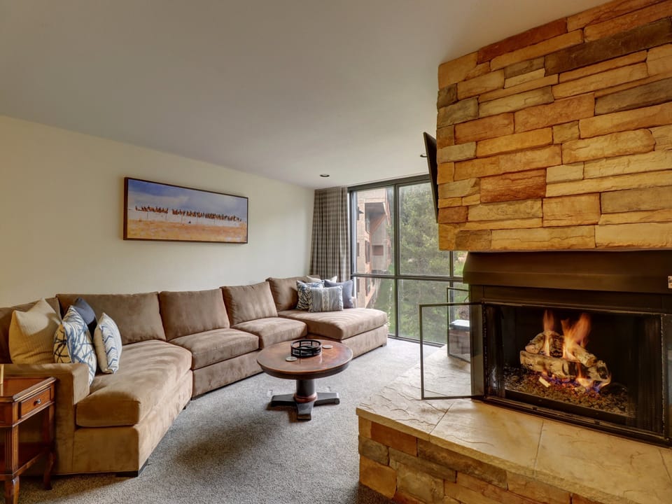 A cozy living room with a large sectional sofa, a wooden coffee table, a stone fireplace with a fire burning, and a large window overlooking the outdoors. A landscape painting hangs on the wall.