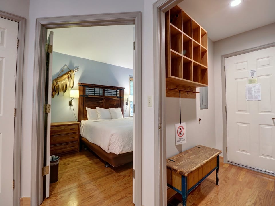 A bedroom with a white bed viewed through an open door on the left, and an entryway with storage cubbies, a bench, and a no-smoking sign on the right.