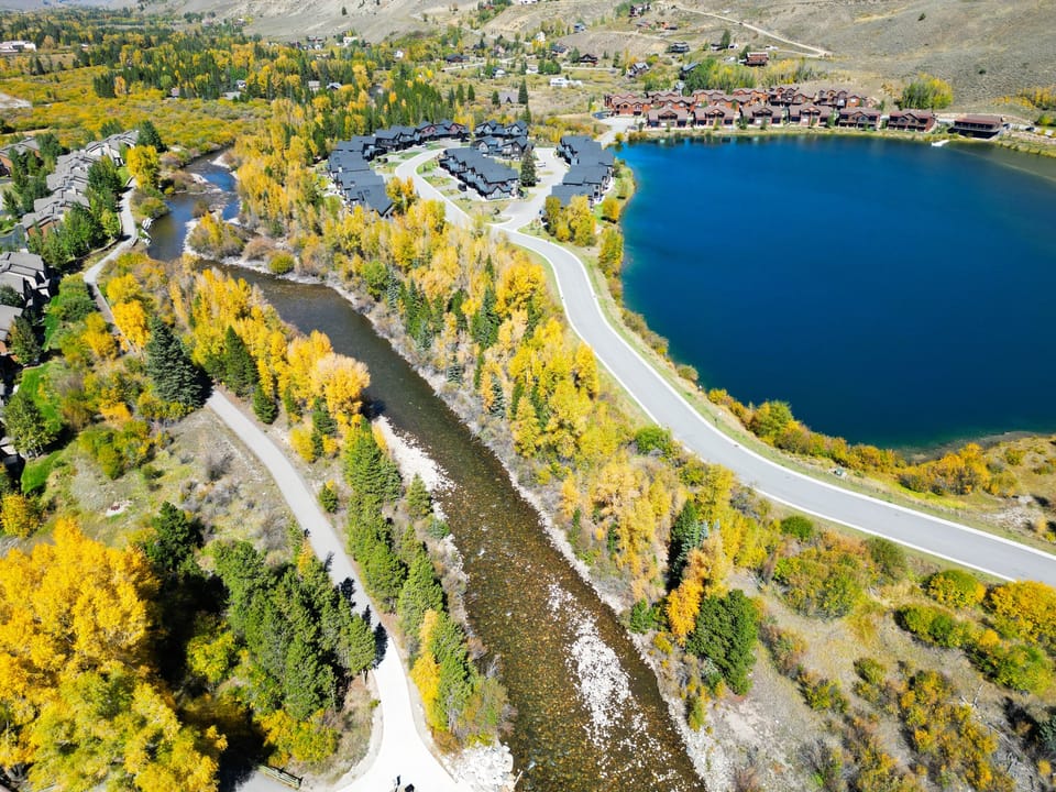 Aerial view of a scenic landscape featuring a clear blue lake, flowing river, surrounding roads, and buildings, all amidst vibrant autumn foliage.