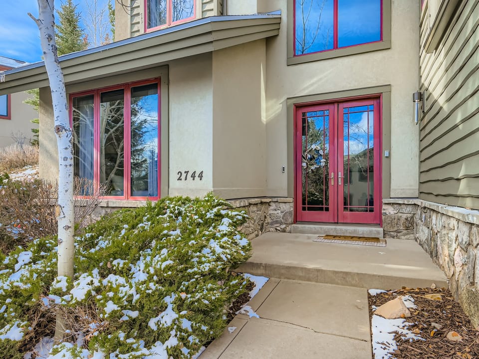 The welcoming entrance features a red-framed double door and number 2744, surrounded by natural stone accents and landscaped greenery dusted with winter snow—a quiet arrival moment in a beautiful mountain setting.