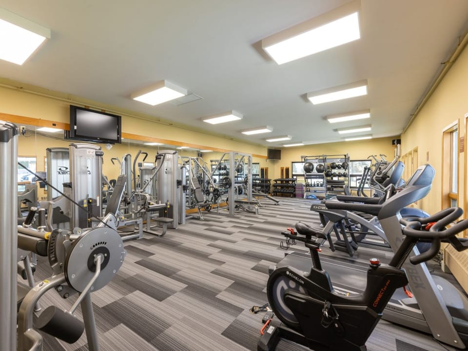 A modern, well-lit gym with various exercise equipment, including treadmills, stationary bikes, weight machines, and free weights, arranged in a spacious area with gray and black flooring.