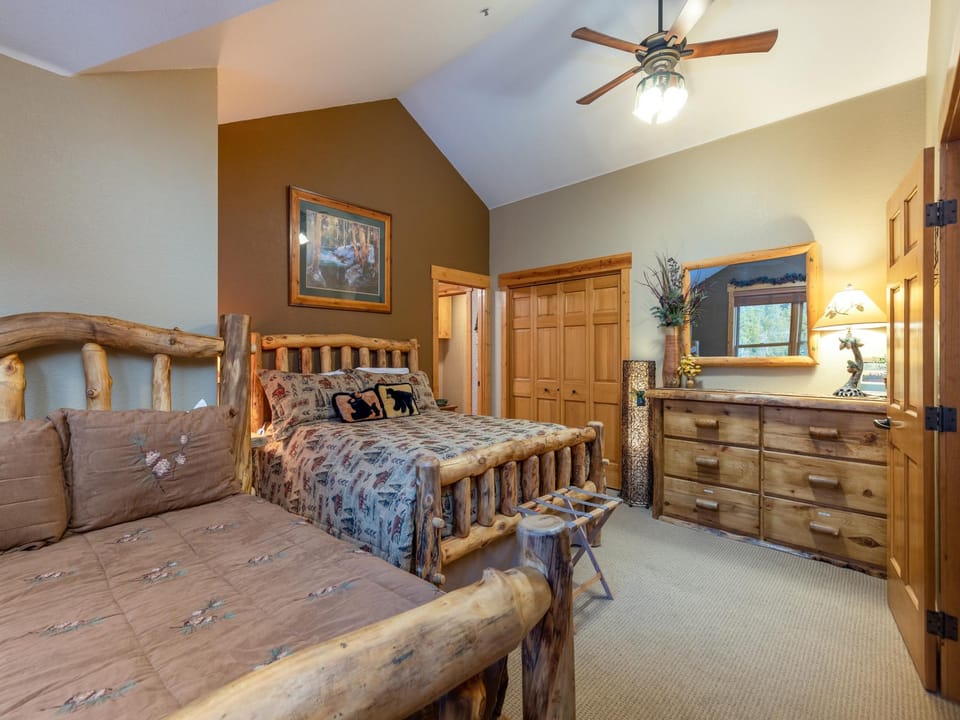 A rustic bedroom with two wooden log frame beds, a dresser with a mirror, and a ceiling fan. The room features earthy tones and outdoor-themed decor, including a nature painting on the wall.
