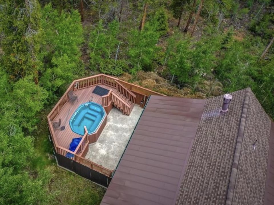 Aerial view of a hexagonal deck attached to a house, featuring a hot tub and seating area, surrounded by lush green trees.
