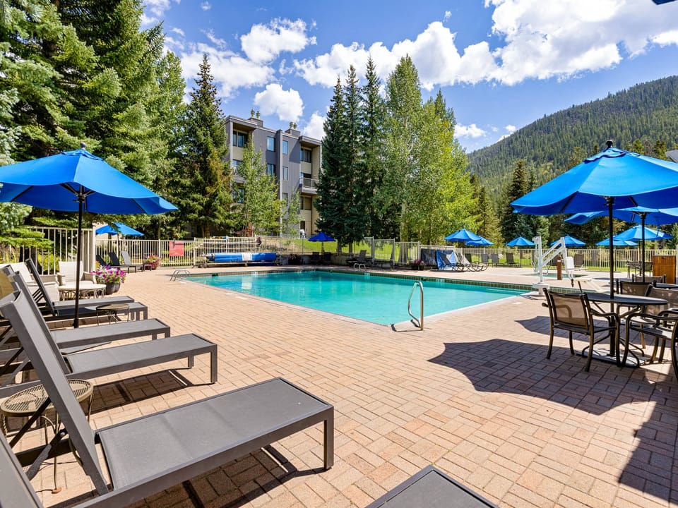 Outdoor swimming pool area with lounge chairs, blue umbrellas, and surrounded by trees and mountains in the background on a sunny day.