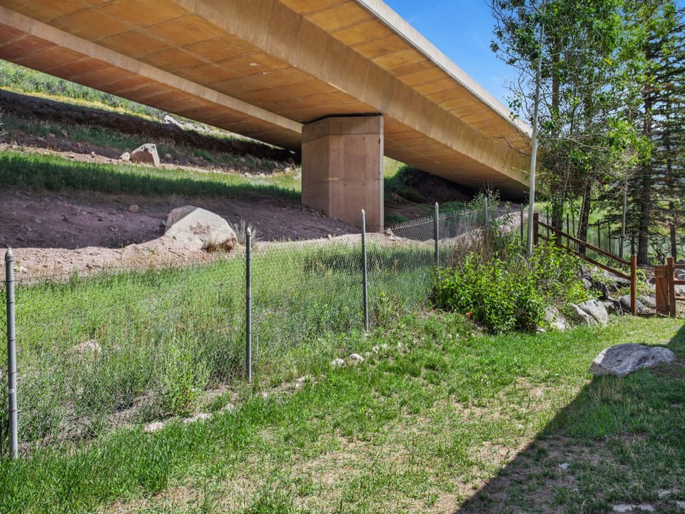A concrete bridge crosses over a grassy area with a chain-link fence and trees on a sunny day.