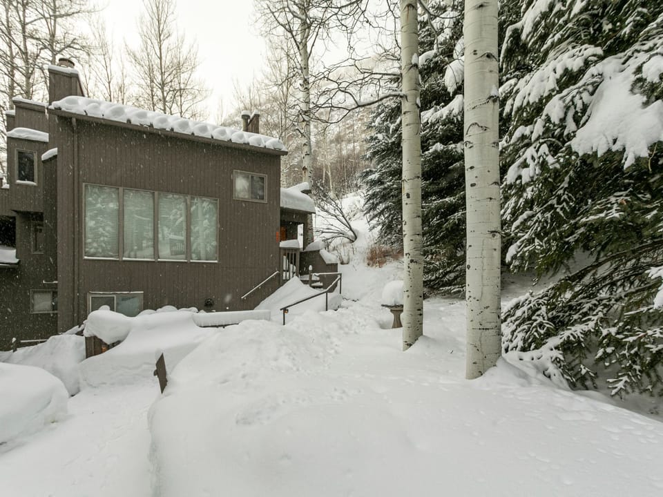 Two-story house surrounded by snow-covered trees and heavy snowfall. Snow accumulates on the roof, ground, and balcony, indicating a cold winter day.