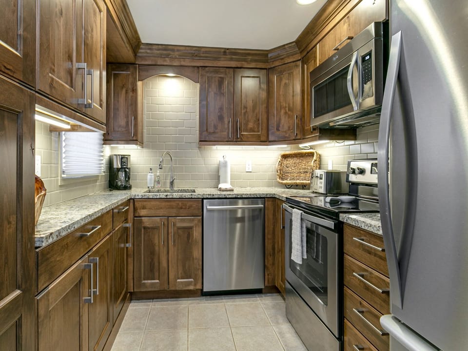 A modern kitchen with wooden cabinets, stainless steel appliances including a refrigerator, microwave, stove, and dishwasher, granite countertops, and a tiled floor and backsplash.