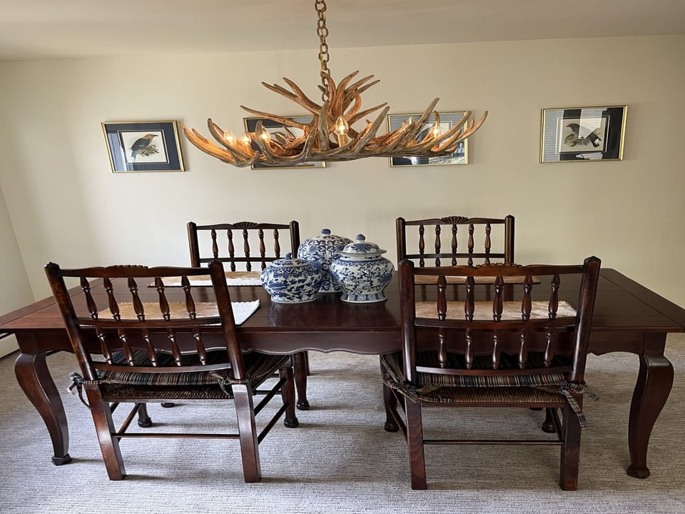 A wooden dining table with six chairs, decorated with blue and white ceramic jars. An antler chandelier hangs above, and three framed pictures are mounted on the wall in the background.